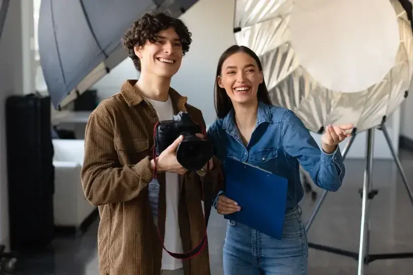 The owners of a photography business—a man holding a camera and a woman holding a clipboard—smile as they prepare for a photo shoot.