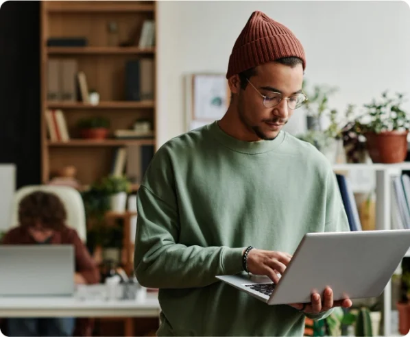 A man in a brown beanie and glasses holds his laptop and types one handed.