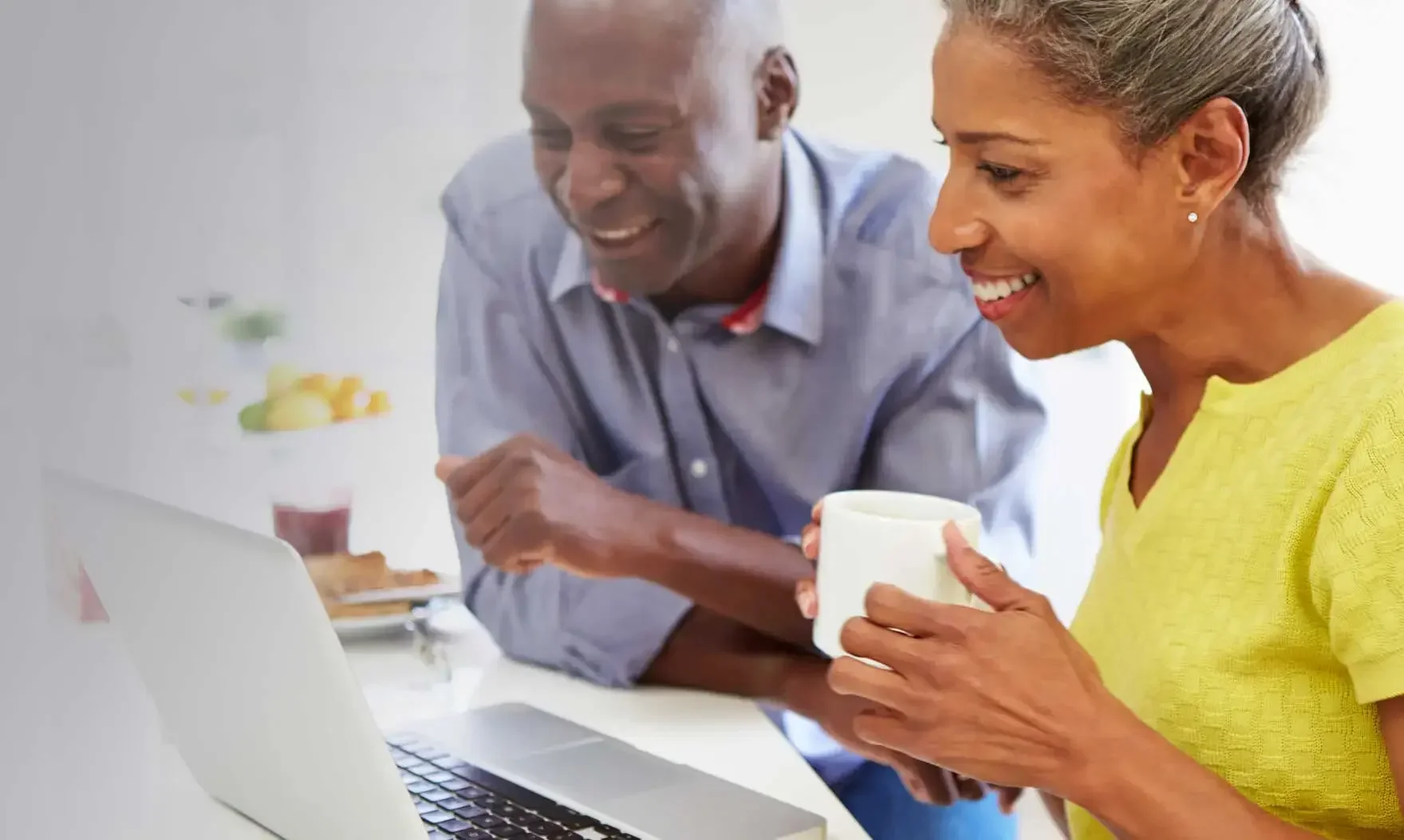 Smiling older couple in a kitchen, looking at a laptop; the woman holds a coffee mug.