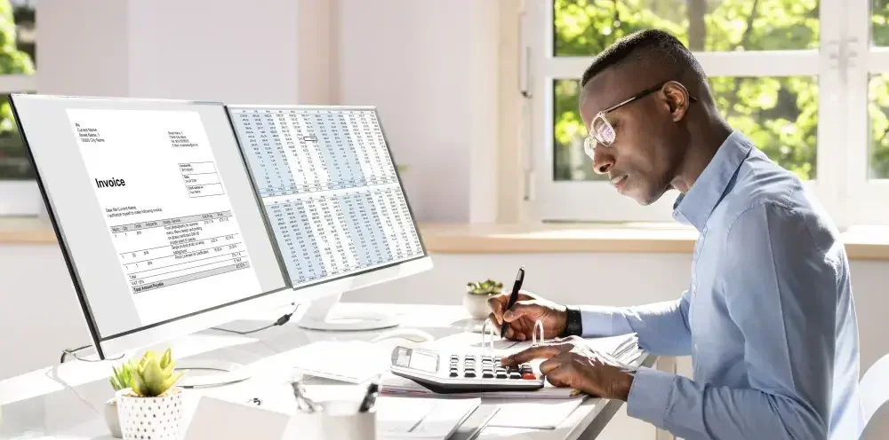 A bookkeeper sits at his desk in front of a dual monitor computer. He reviews invoices and enters data into a spreadsheet while checking his work with a calculator.