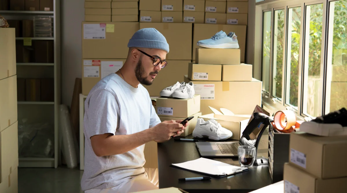 A man sits at a desk and packs a customer order. He's surrounded by shoeboxes and sneakers.