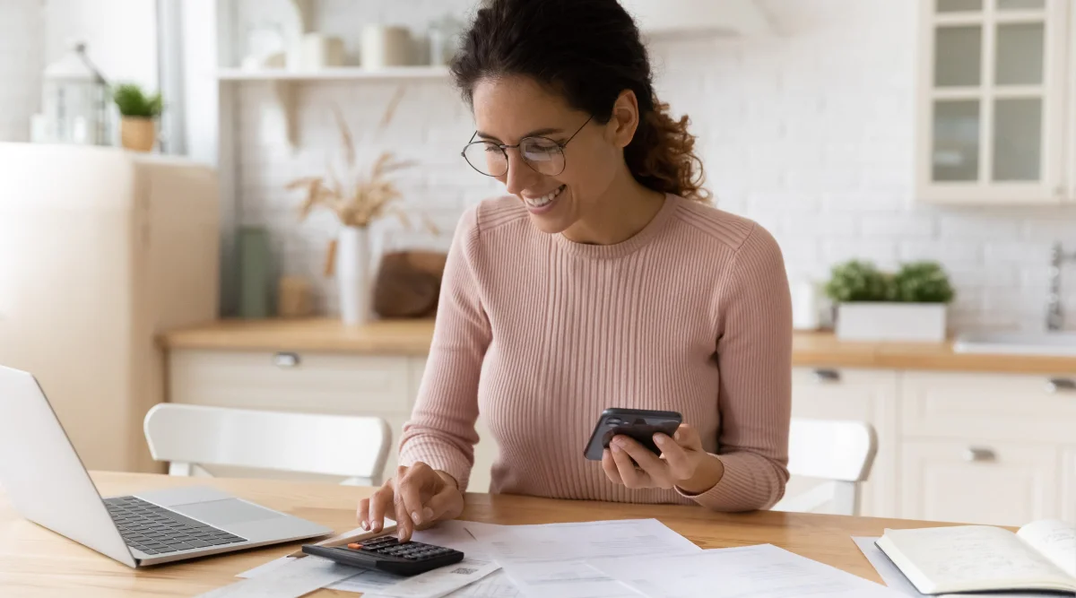 A woman calculates figures on a calculator, smiling as she manages paperwork at her kitchen table