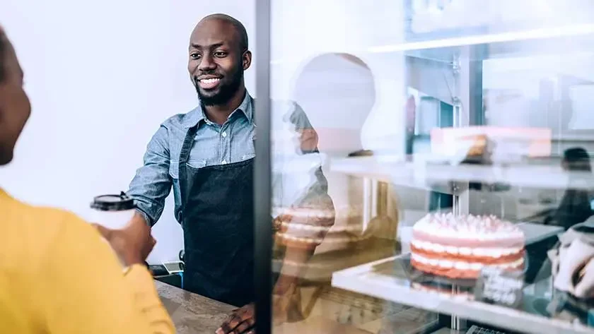 Black man in a denim button down and a black apron giving takeaway coffee to customer in a yellow shirt confectionery shop. Cakes and desserts in the background