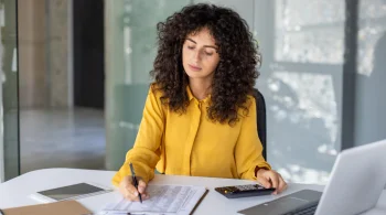 Woman in yellow writing with a calculator and laptop