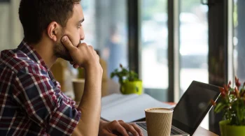 Man in checked shirt thinking with laptop and coffee