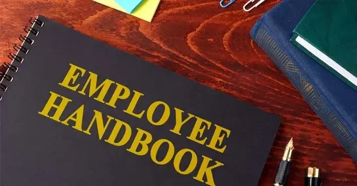 A book whose cover reads "Employee Handbook"sitting on a table surrounded by office supplies