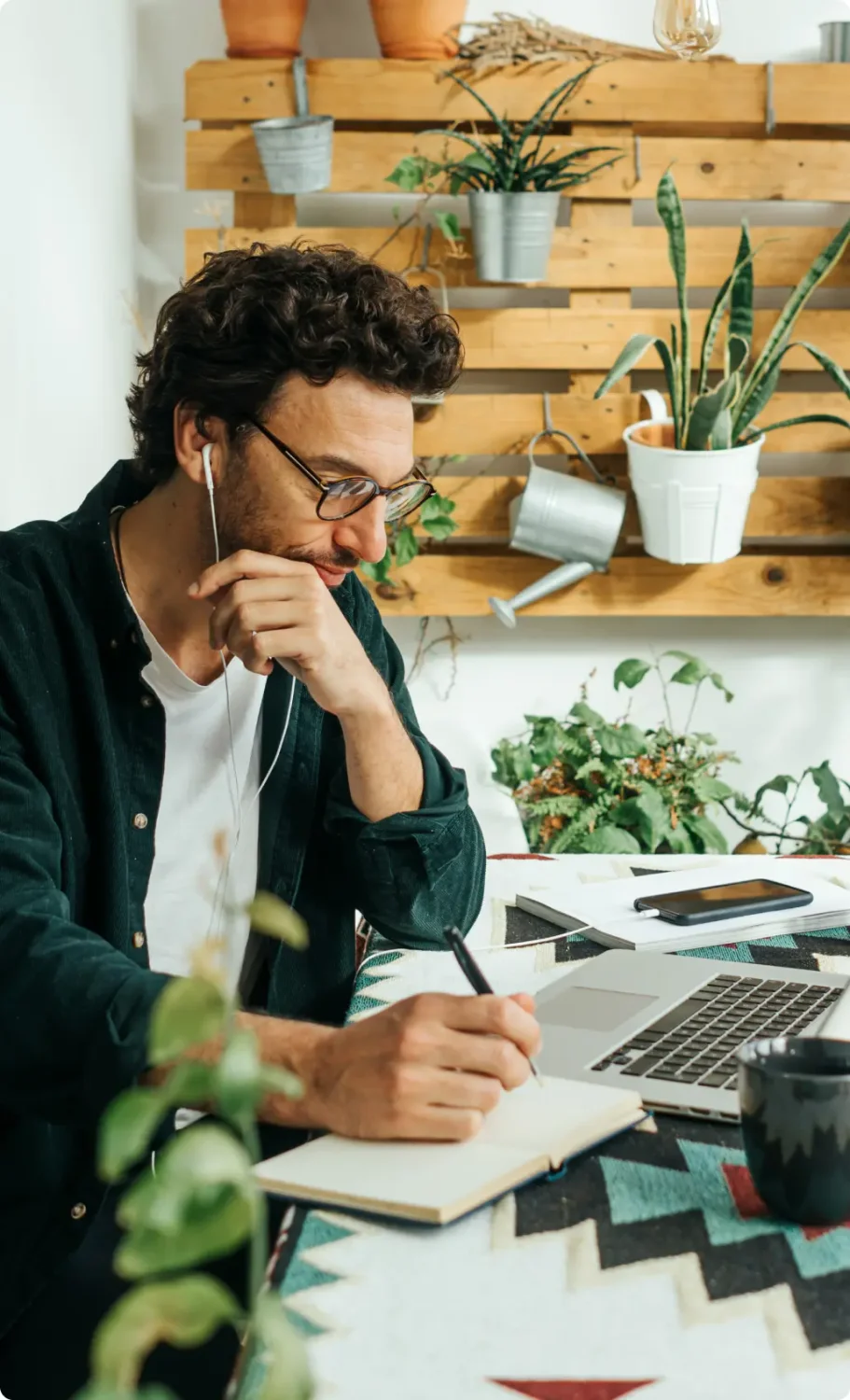 Man wearing glasses and earbuds writes in his journal next to his laptop.