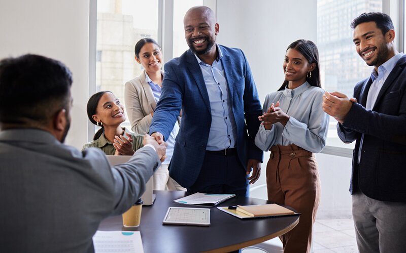 An image of 6 people in a meeting room while 2 of them are shaking hands and the rest are clapping.