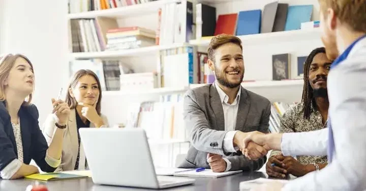 Five businesspeople sit around a conference table, two men shaking hands