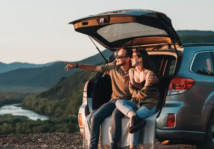 A man and a woman sit in the back of their open car, as they look out onto a sunset. The man points and they both smile.