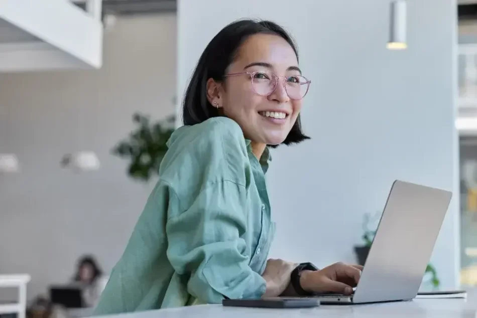 A woman seated at a desk in an office smiles as she looks up from her laptop, on which she has been researching NY LLCs.