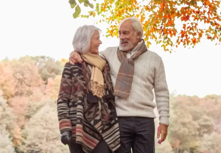 An older man and woman smile into each other's eyes as they walk through orange and red leaves in the fall.