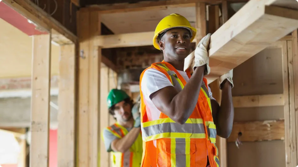 Two men wearing yellow hard hats and construction safety vests carrying a long piece of lumber.