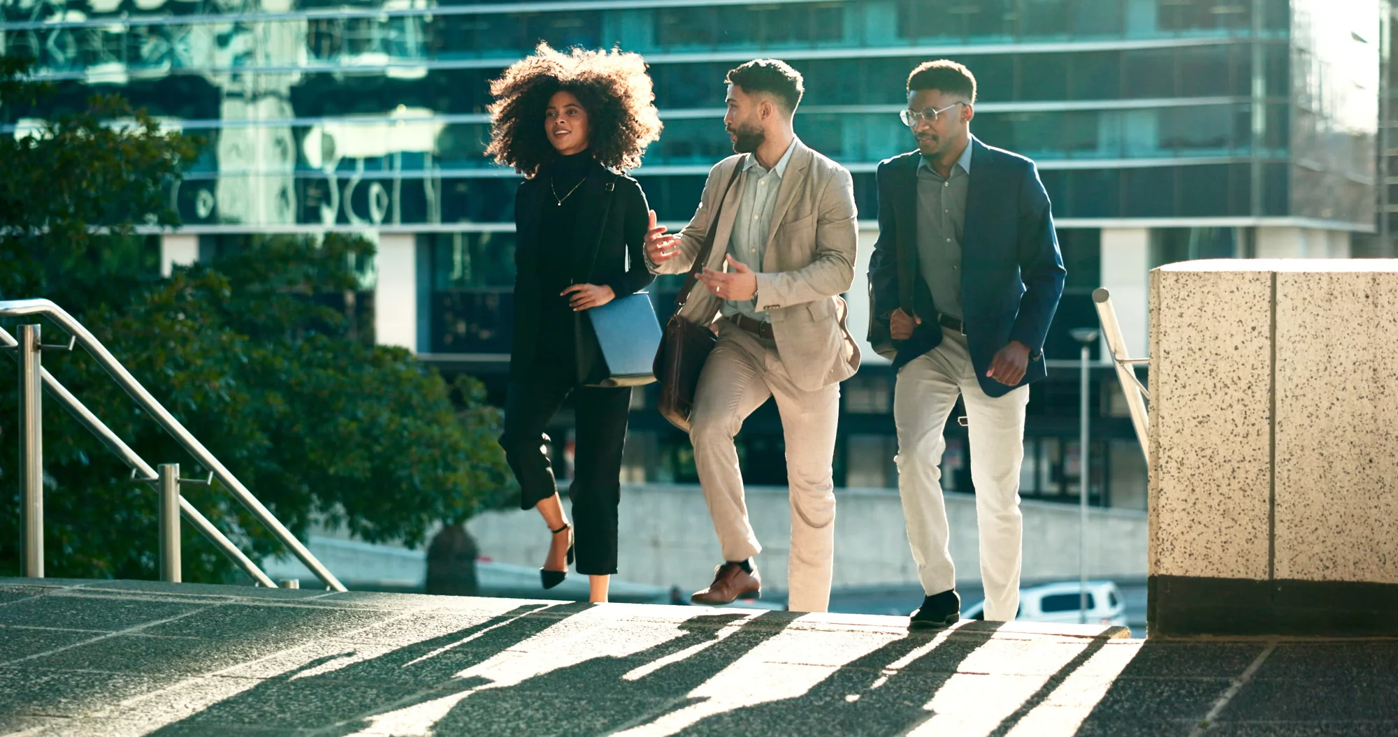 Three professionally dressed individuals walking up outdoor steps in an urban setting, engaged in conversation with a modern glass building in the background.