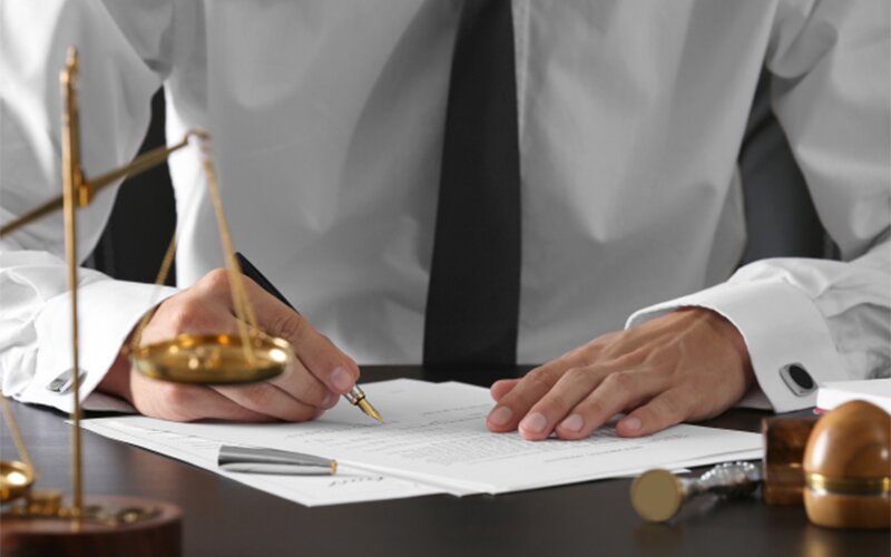 An image of a man signing a document with a balancing scale and gavel on the table.