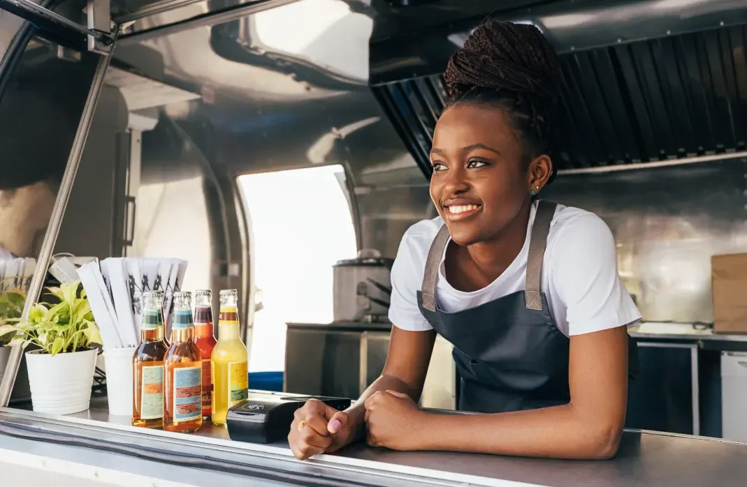 A black woman wearing an apron leaning on the opening of a foodtruck.