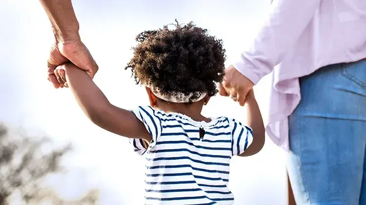 A toddler walks while holding the hands of two adults
