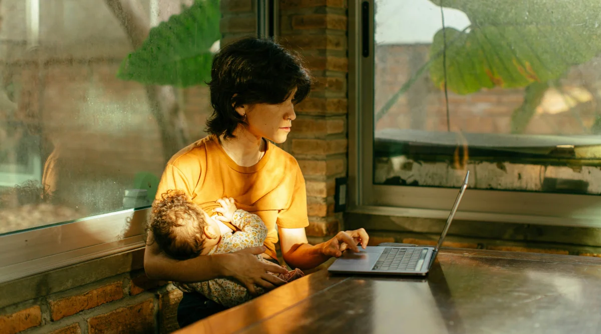 A woman holds a baby in one arm and uses a laptop with the other.
