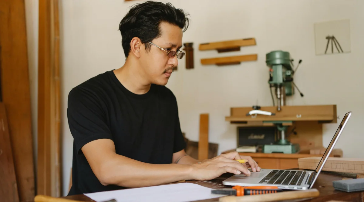 A man in a black shirt works on a laptop in a workshop filled with woodworking tools