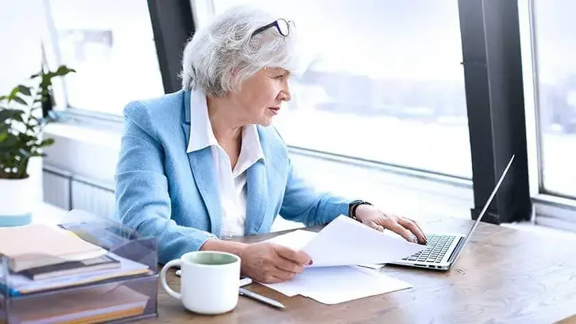 A woman sits at her desk looking at papers and taking notes on her laptop.