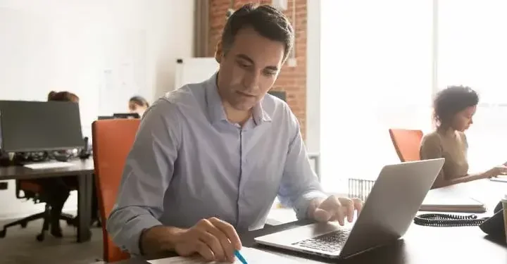 Man in foreground of an office environment works on his laptop while checking against his notes on paper.
