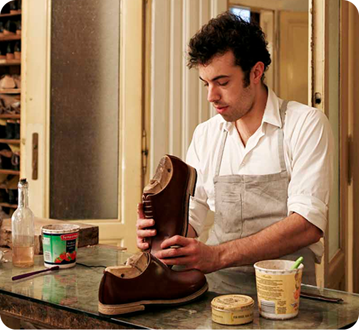Male cobbler in overalls works on a pair of shoes in his shop