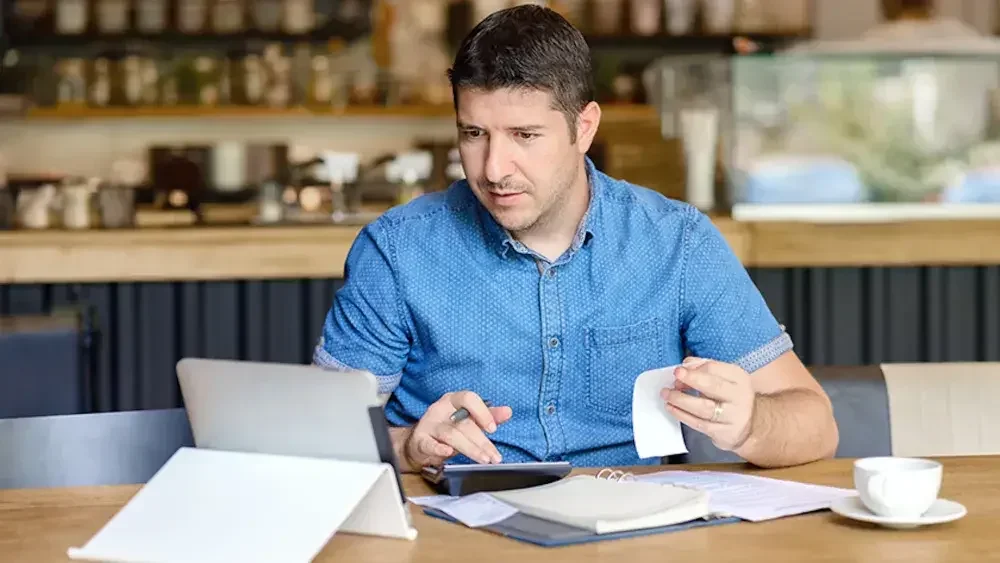 A cafe owner sits at a table looking at his tablet and using a calculator.