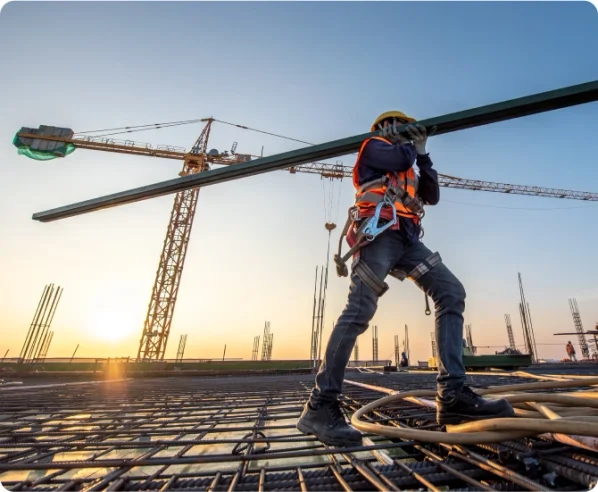 A construction worker in a high visibility vest carries a metal beam on his shoulder, through an jobsite.