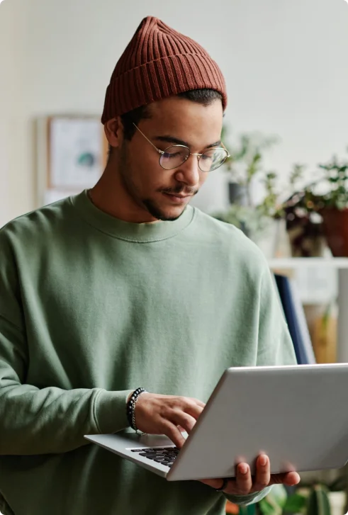 A man in a brown beanie and glasses holds his laptop and types one handed.