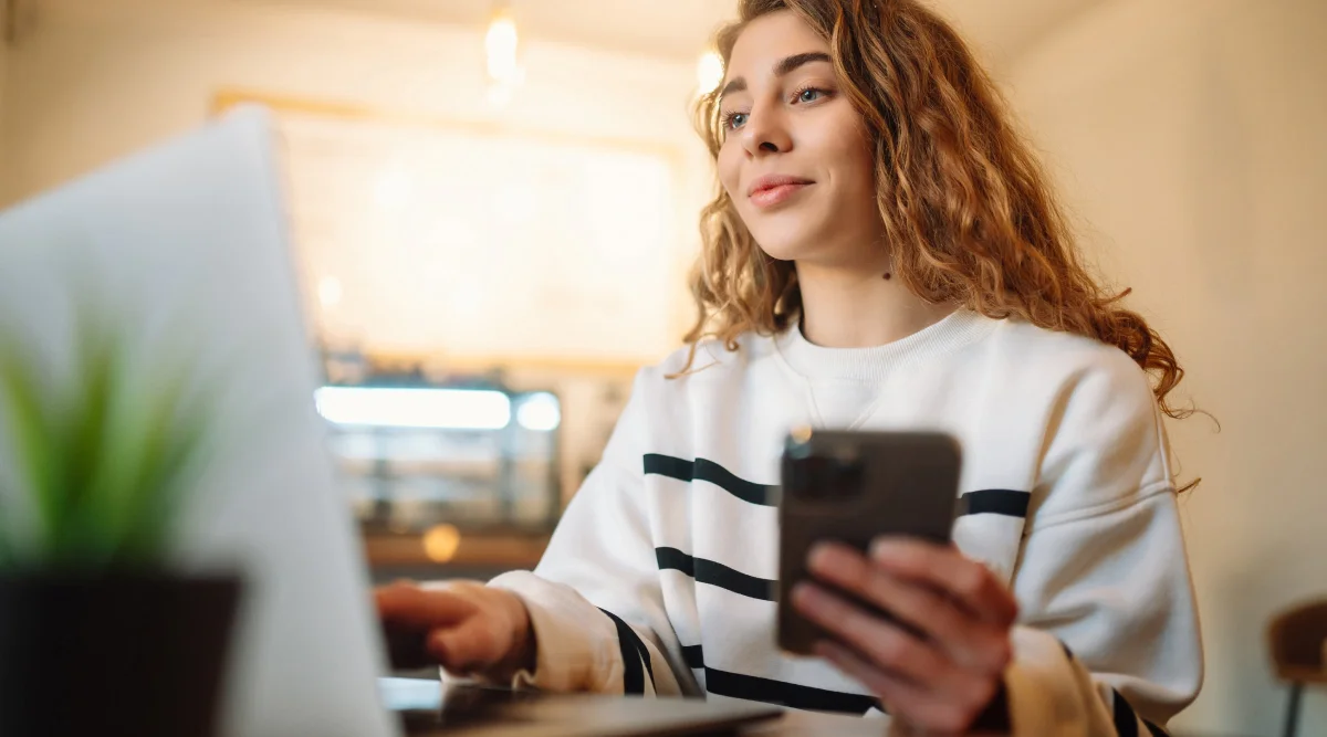 A woman smiles as she checks her phone while reading her laptop