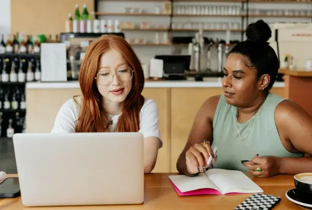 two women study a laptop while one takes notes on paper.