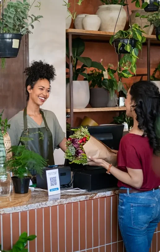 Smiling african woman botanist selling flowers and plants to a customer while standing in flower shop. Happy black young woman entrepreneur standing behind counter wearing apron in plant store selling fresh flowers to client. Young latin girl buy a fresh bouquet from florist.