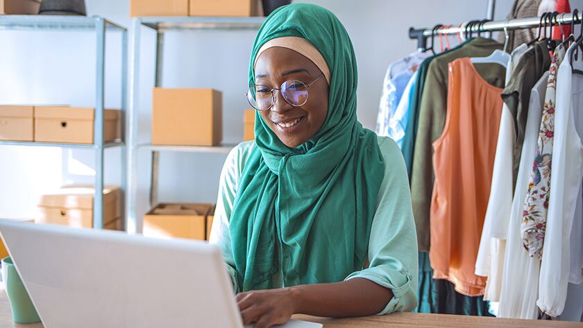 A clothing designer wearing a headscarf sits in her shop with a rack of clothes and boxes stacked behind her. She is forming her single-member LLC online.