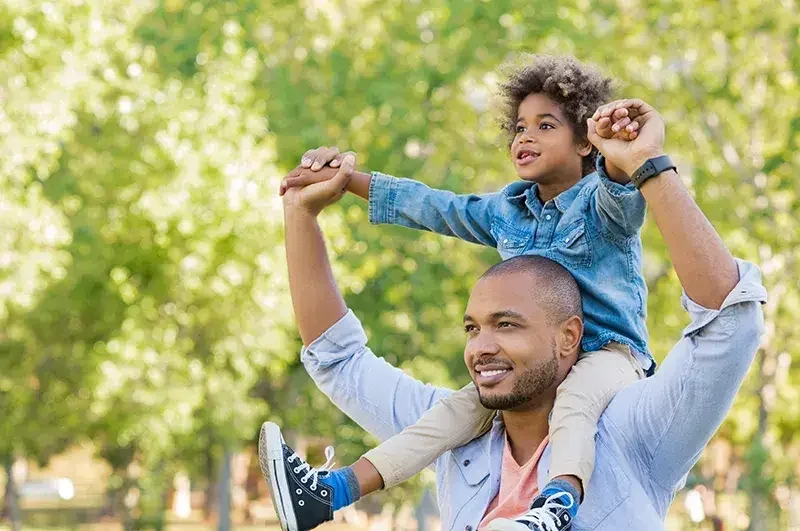 An image of a man carrying a kid on his shoulders in a park.