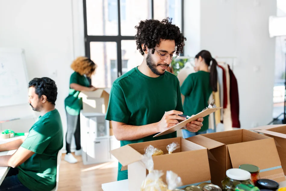 Man wearing a green shirt smiling and looking down at a clipboard surrounded by other employees and cardboard boxes.