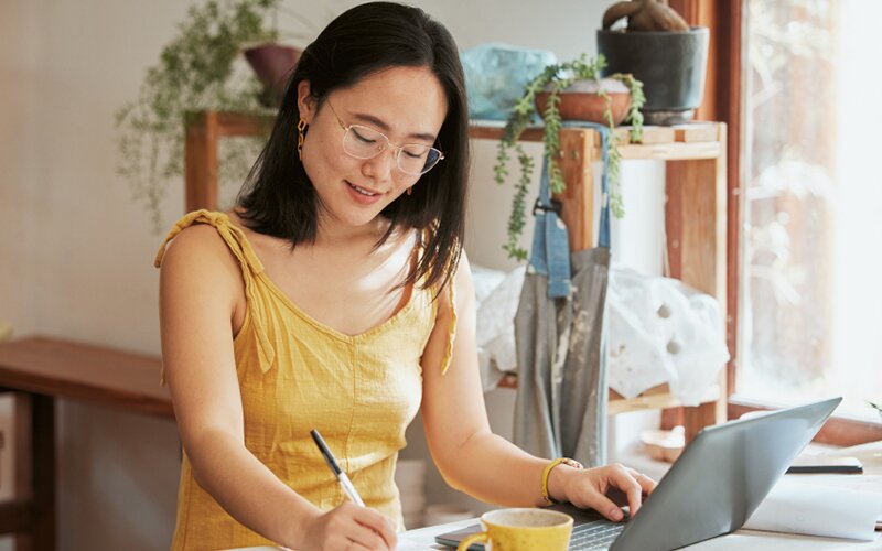 An image of a woman making notes while working on her laptop in her home office.