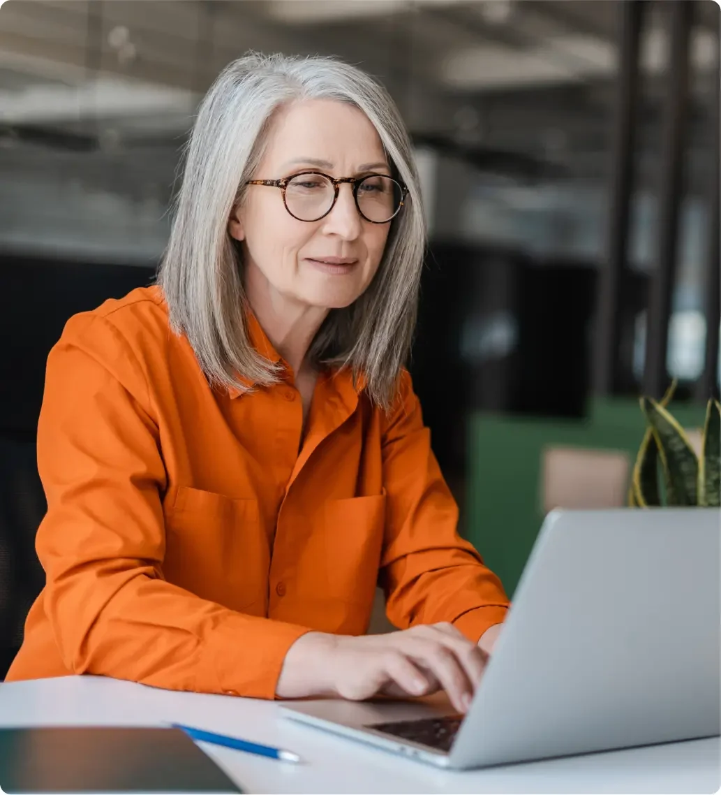 Older White woman with grey hair wearing glasses and a long sleeved orange shirt working on her LegalZoom corporate dissolution on her laptop.