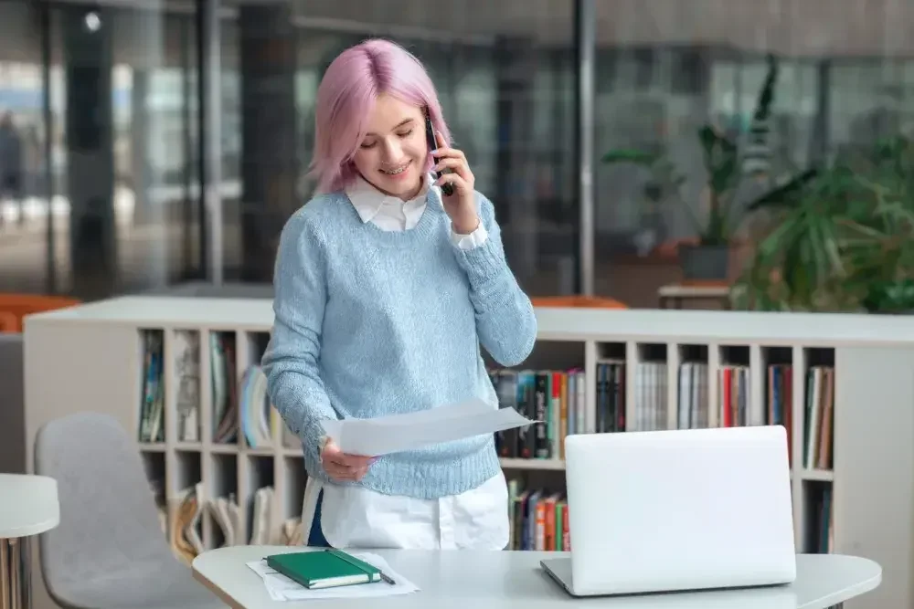 A woman with pink hair holds a cell phone with one hand and a BOIR fact sheet in the other hand as she stands in front of her desk in her office.