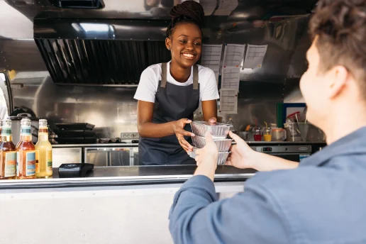 Black woman with her hair pulled back with a headband in an apron working at food truck giving takeaway packaged food to customer.