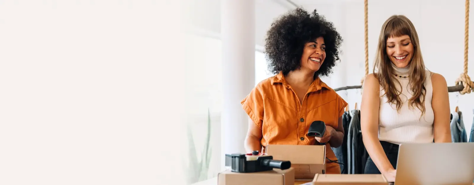 Black woman with an afro wearing a orange romper talking to her business partner, a blonde woman wearing a white sleeveless shirt talking about setting up their registered agent with LegalZoom as they mail packages to customers.
