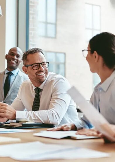 A man wearing glasses and a white button-up shirt and tie is smiling at a brunette woman wearing glasses in a conference room. In the background is smiling as they discuss how to incorporate their business.