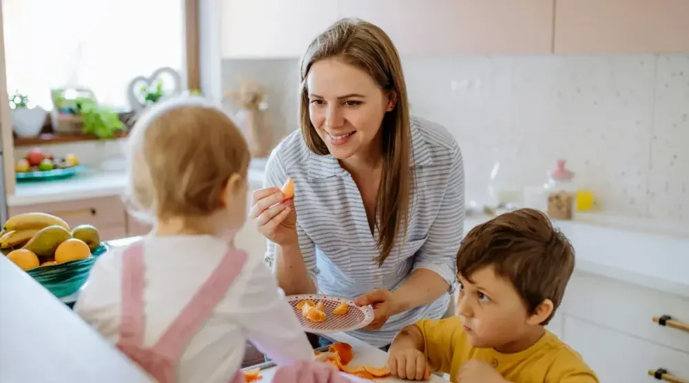 An adult stands in the kitchen feeding. two children.