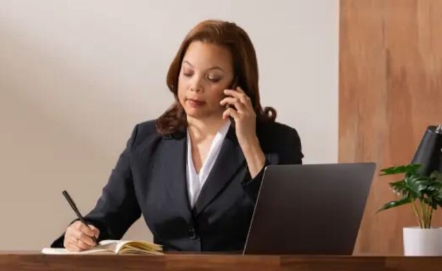 a woman wearing a suit behind her desk writes a note and speaks on the phone