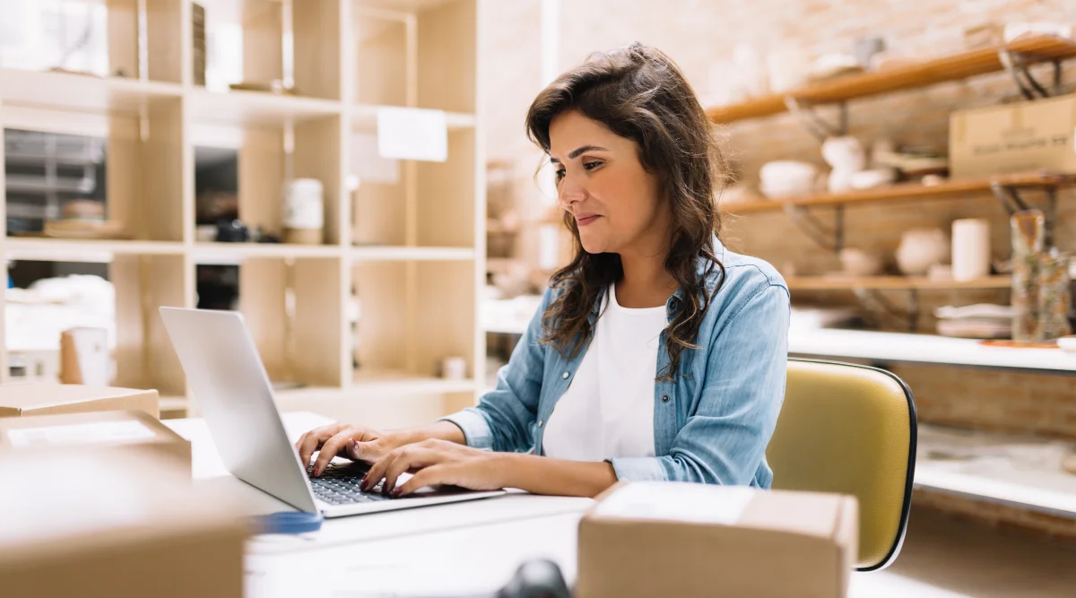 A woman sits in her pottery shop desk typing on her laptop.