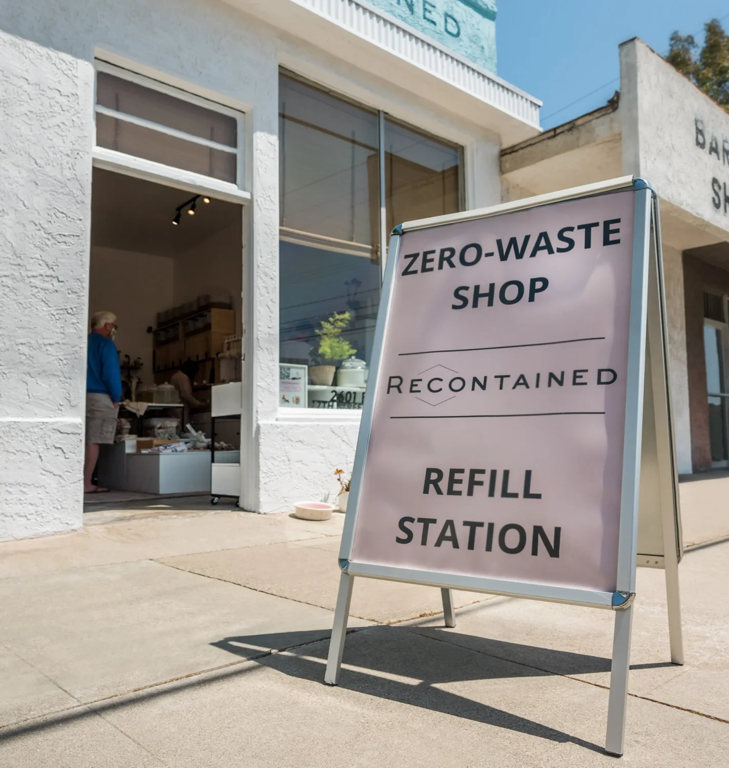 The entrance to a zero-waste shop called 'Recontained' with a sign outside that reads 'Zero-Waste Shop Recontained Refill Station' and a person visible inside the shop.