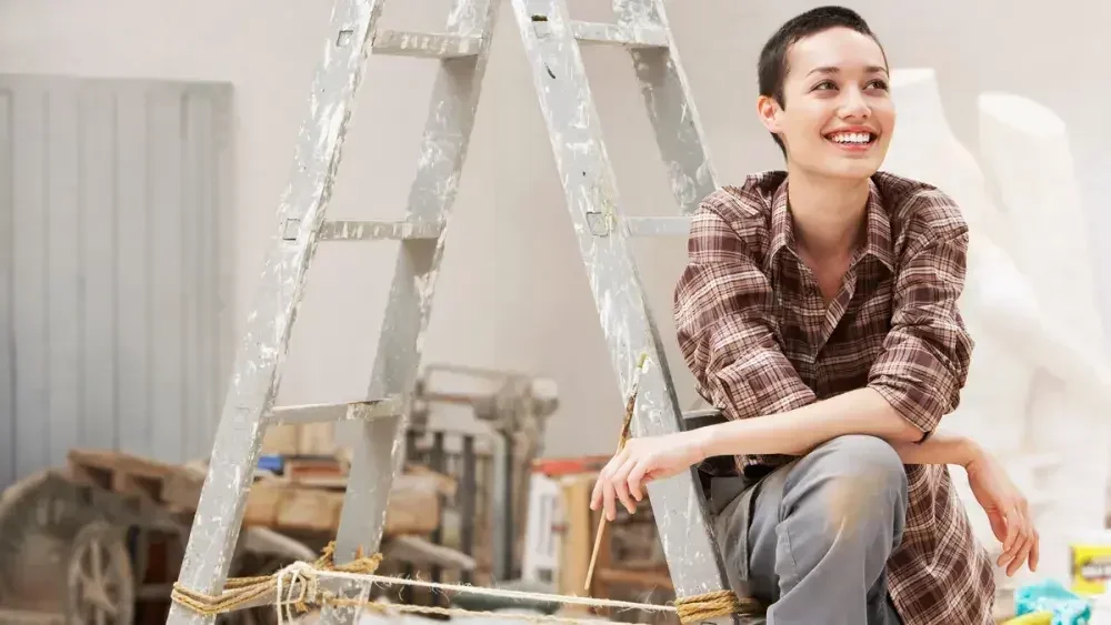 A woman holding a paintbrush takes a break sitting on a ladder.