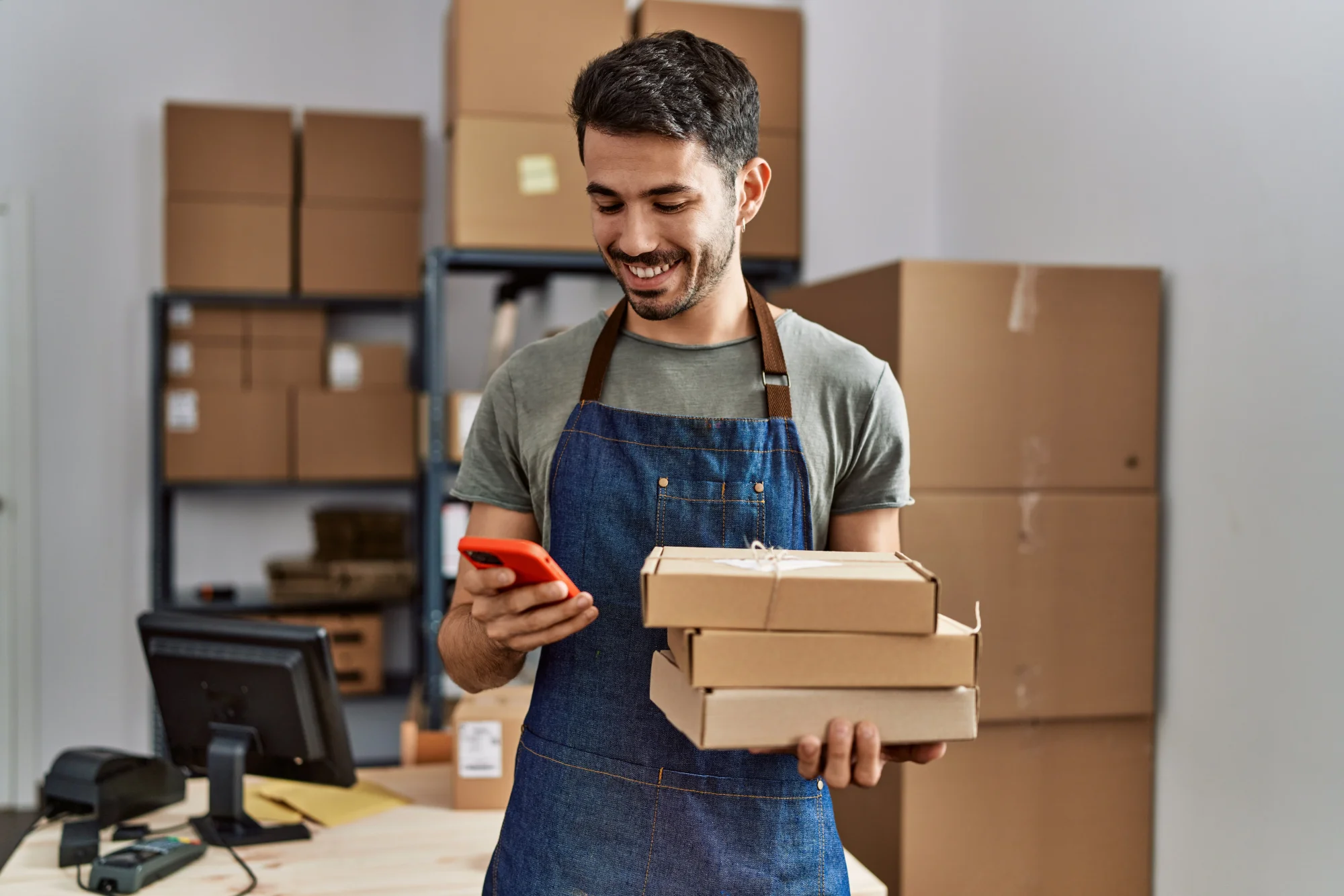 Smiling young man wearing a gray shirt and denim apron standing in a warehouse with boxes in the background looking at his cell phone in one hand and holding packages in the other.