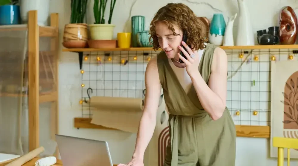 A woman talks on her cellphone while looking up something on her laptop in her ceramics studio.