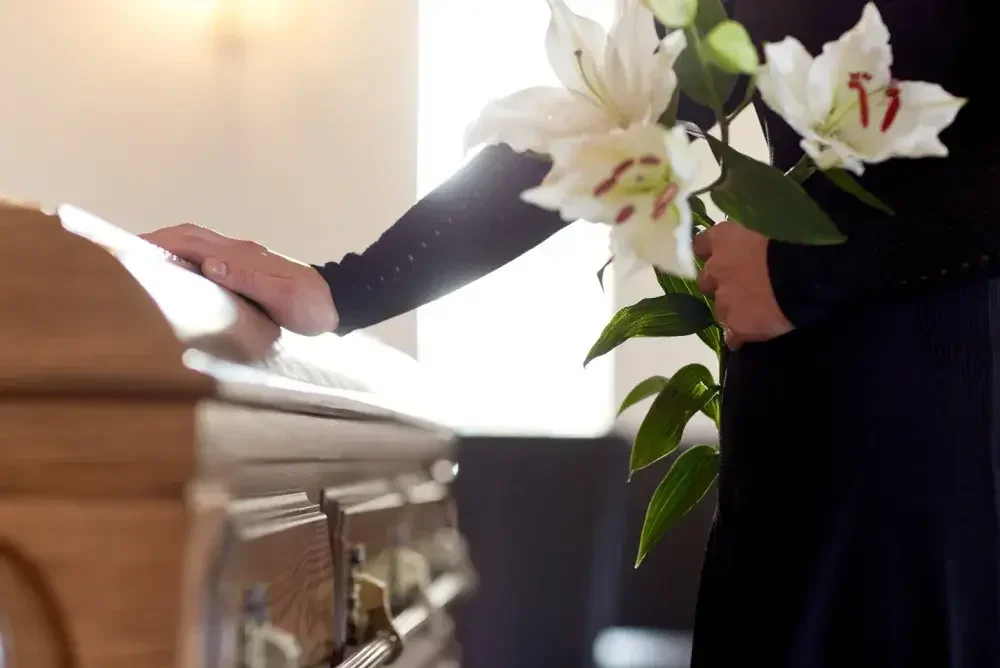 A person in mourning places flowers on top of a coffin. The medical examiner or funeral director will ensure all known and necessary information is included on the death certificate before signing.