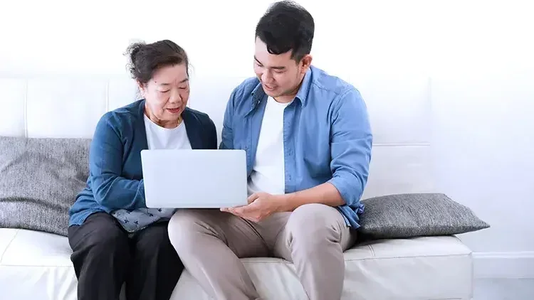 A man and his mother sit on a bed and look at a laptop and understanding how to make a do-it-yourself living trust.